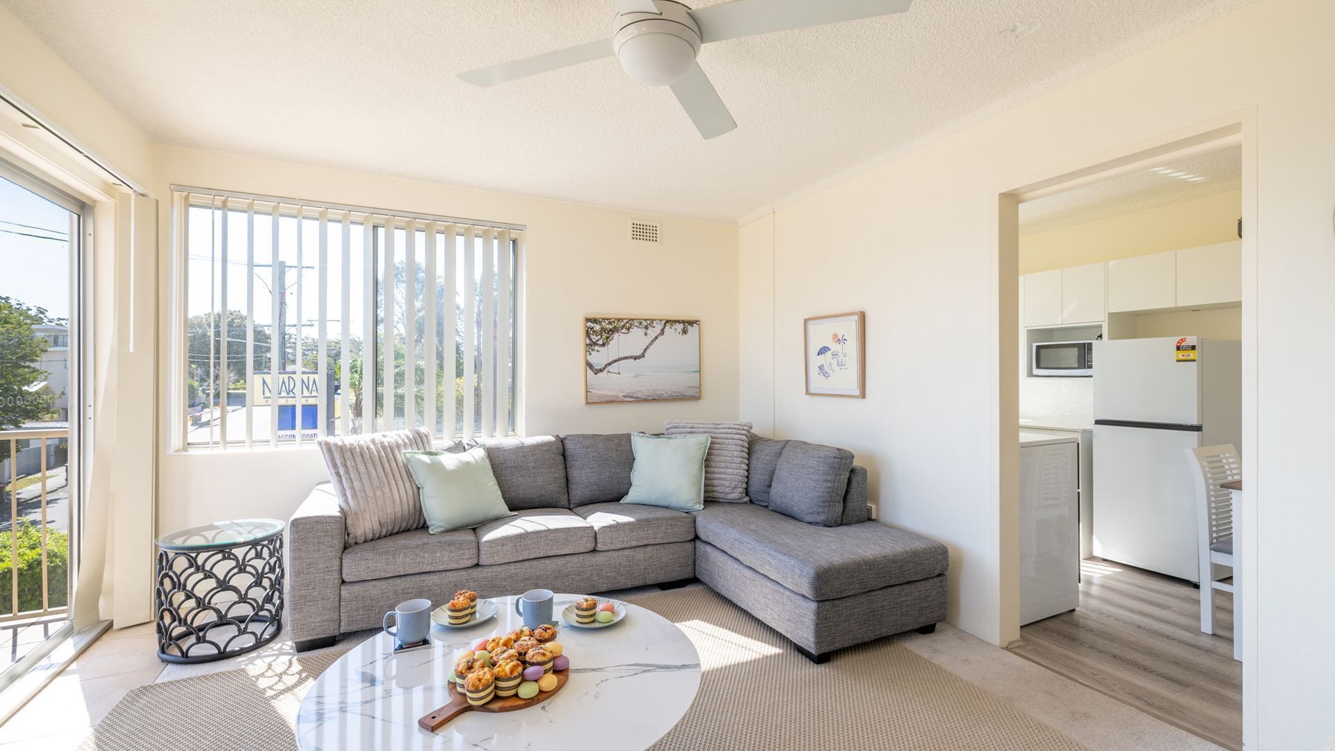 A modern living room with a grey sectional sofa, a round coffee table with pastries, a ceiling fan, and a view of the outside through large windows.