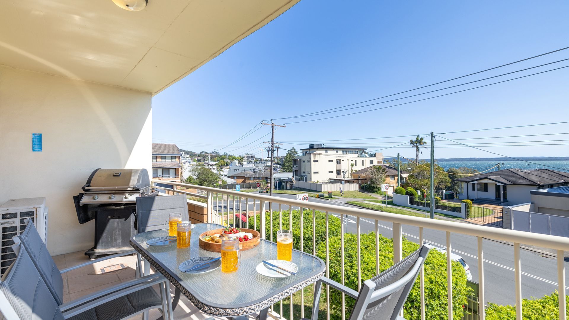 A balcony with a grill, dining table, and chairs, overlooking a residential area with houses and a body of water.