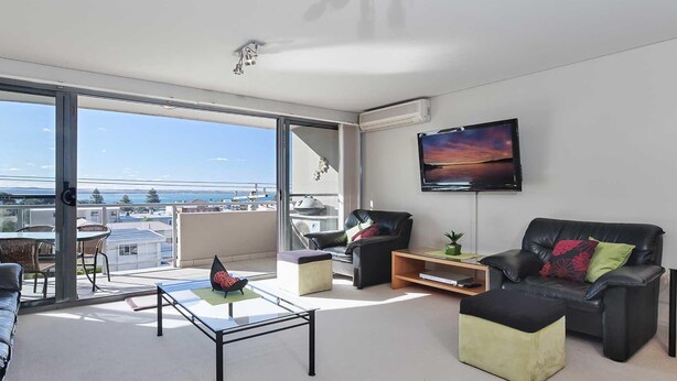 A modern living room with a large sliding glass door leading to a balcony, a flat-screen TV mounted on the wall, and a comfortable seating area with a sofa and chairs. - Bayview Apartments, 11,42 Stockton Street - Three bedroom unit In the heart of town with air con, lift and secure parking (Nelson Bay)