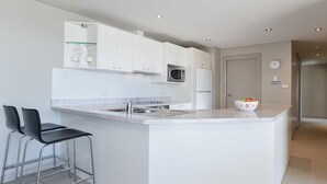 A modern kitchen with white cabinetry, a stainless steel sink, a microwave, and a bowl of fruit on the counter. There are two black bar stools and a glass shelf above the sink. - Bayview Apartments, 11,42 Stockton Street - Three bedroom unit In the heart of town with air con, lift and secure parking (Nelson Bay)