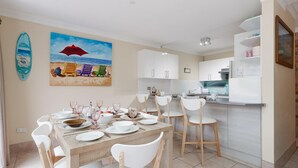 Zona pranzo con tavolo apparecchiato per sei, sedie bianche e un quadro raffigurante sedie da spiaggia.