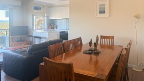 A spacious living area with a wooden dining table, leather sofa, and a view of the kitchen and balcony. - Adriana, 9/83 Ronald Avenue - fabulous views & aircon (Shoal Bay)