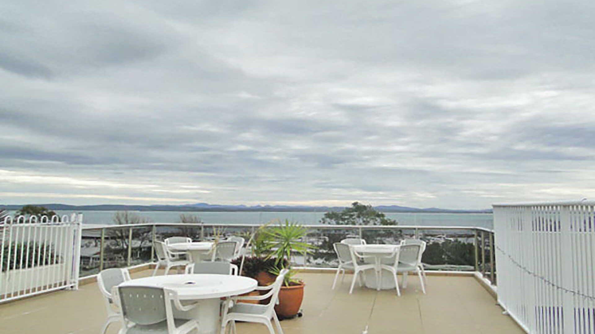 Une terrasse avec des tables et des chaises blanches, des plantes en pot et une vue sur la mer et les montagnes lointaines.