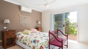 Une chambre avec un lit, deux tables de chevet, une décoration murale en macramé, un ventilateur de plafond et une grande porte-fenêtre coulissante donnant sur un espace extérieur.