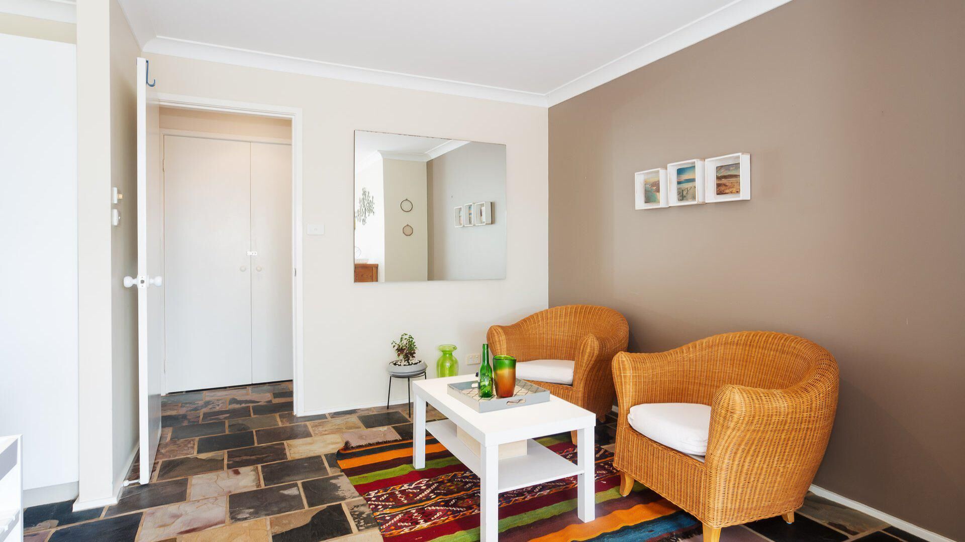 A room with a white door, a mirror, two wicker chairs, a small table with a colorful rug, and a brown wall with three framed pictures.