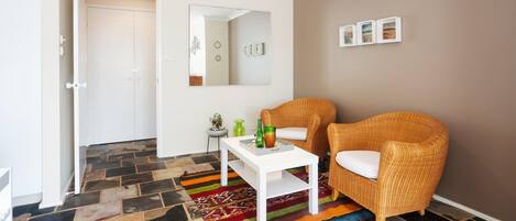 A room with a white door, a mirror, two wicker chairs, a small table with a colorful rug, and a brown wall with three framed pictures.