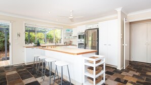 Une cuisine moderne dotée d’un grand îlot central, de meubles blancs et d’un four encastré. On y trouve trois tabourets de bar et un ventilateur de plafond.