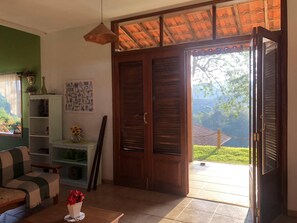 A cozy living room with a sofa, a coffee table with a vase of flowers, a bookshelf, and a sliding door leading to a balcony with a view of greenery. - Beautiful country house with swimming pool and panoramic views 1 hour from São Paulo (Extrema)
