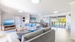 A modern living room with a flat-screen TV, grey sofas, a dining area, and a sliding glass door leading to a pool.