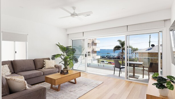 A modern living room with a grey sofa, a wooden coffee table, and a large sliding glass door leading to a balcony with a view of the sea and parked cars. - Hideaway 1, 3 Lillian St -  stunning water views (Shoal Bay)