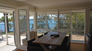 A covered patio with a wooden table and chairs, large windows offering a view of the water and trees. - Wanda Magic, 19 Randall Drive - beautiful water front house (Salamander Bay)