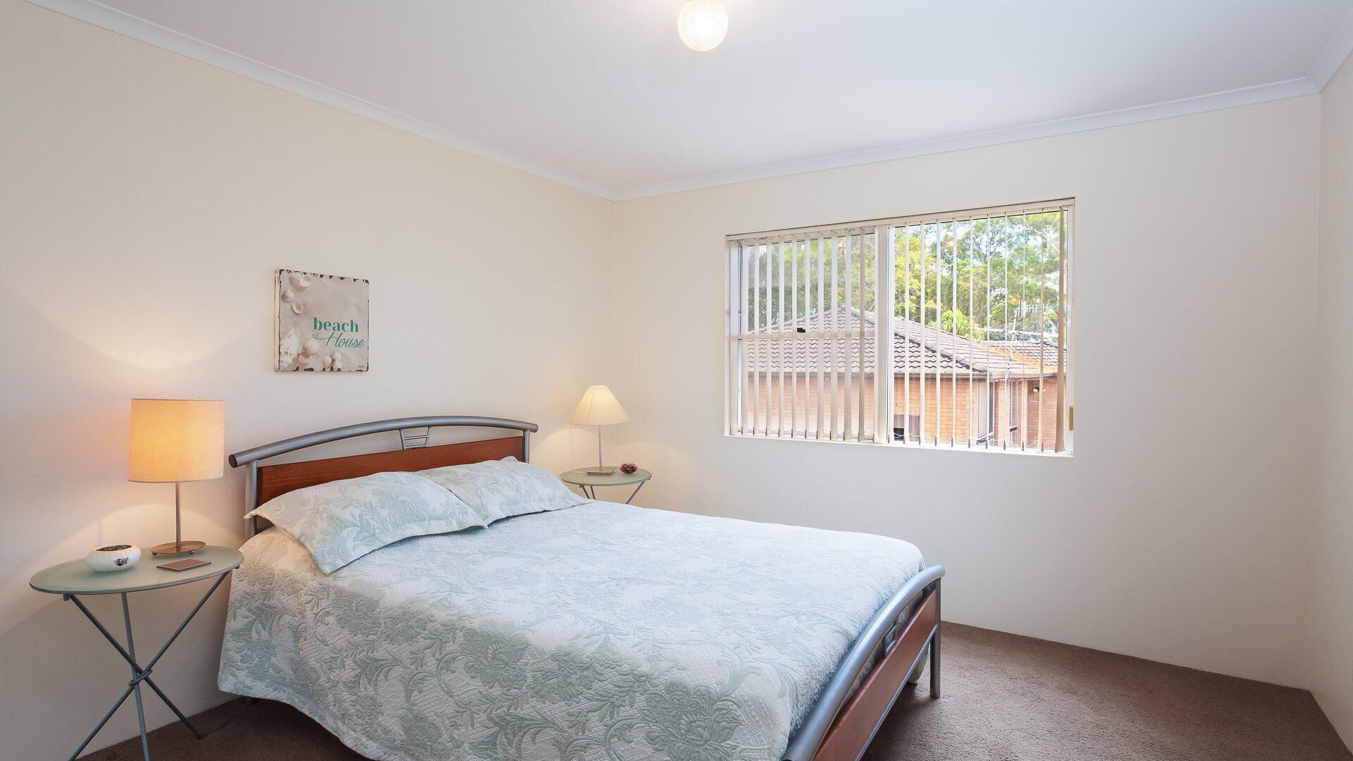 A bedroom with a bed, two bedside tables, a window with blinds, and a wall-mounted sign.