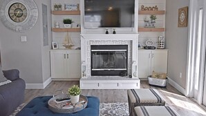 Ein Wohnzimmer mit Kamin, einem Fernseher darüber und einer Couch. Regale mit Dekorationselementen und eine Uhr sind an der Wand angebracht.
