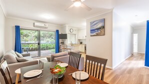 A modern living room with a dining area, a kitchen, and a large sliding glass door leading to a garden.