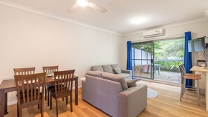 A compact living space with a dining table, wooden chairs, a grey sofa, and a small kitchen area with a TV and a kettle.
