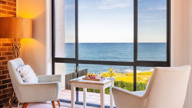 Une pièce avec une grande fenêtre donnant sur la mer, deux chaises, une petite table avec un verre de vin et un mur en briques.