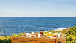 Une table en bois, dressée avec des aliments pour le petit-déjeuner et une cafetière, donnant sur l’océan.