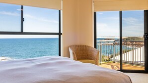 Une chambre dotée d’un grand lit, d’un fauteuil en osier et d’un balcon offrant une vue sur la mer et la côte rocheuse.