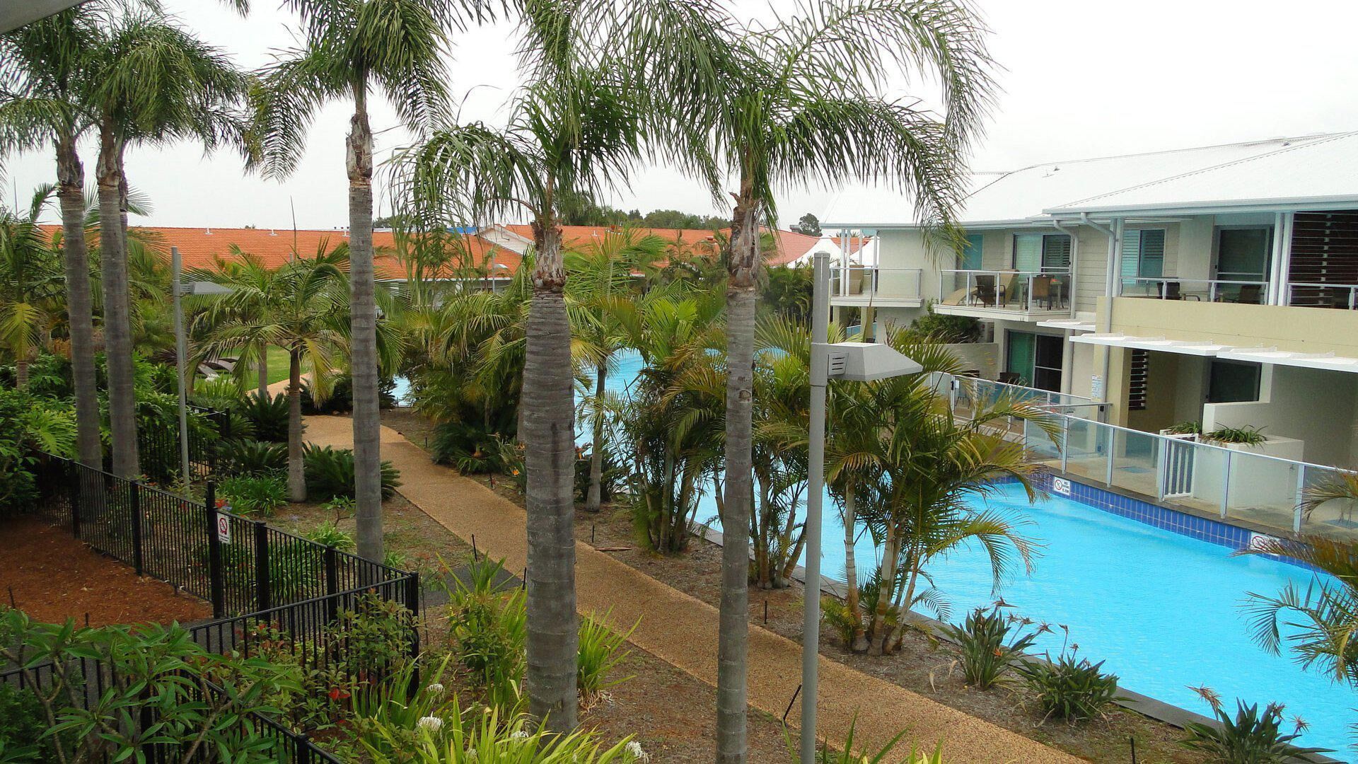 Un hôtel doté d’une piscine entourée de palmiers, d’un jardin clos et d’un bâtiment avec de nombreux balcons.
