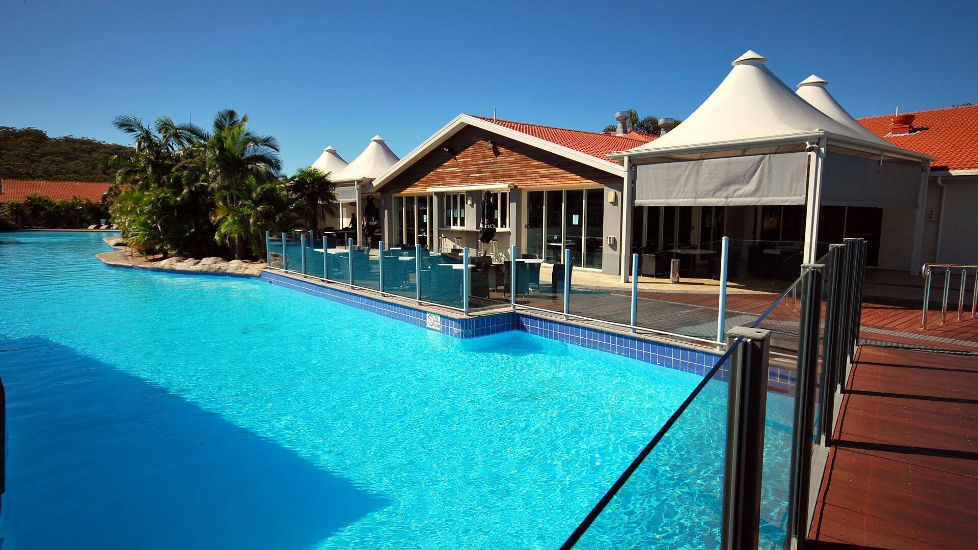 A swimming pool with clear blue water, surrounded by a wooden deck and a covered area with a red roof.