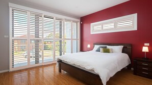 A bedroom with a bed, a nightstand, a lamp, and a window with blinds. - 7 Judith Street - Stunning duplex with ducted air (Corlette)
