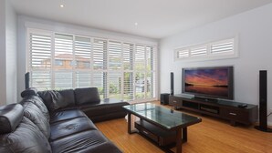 A modern living room with a leather sofa, a glass coffee table, a flat-screen TV, and a large window with blinds. - 7 Judith Street - Stunning duplex with ducted air (Corlette)