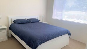 A neatly made bed with a blue bedspread and pillows, a white nightstand with a lamp, and a window with blinds.