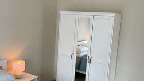Un armari blanc amb dues portes i un mirall, una tauleta de nit amb una làmpada, i un llit amb roba de llit blanca.