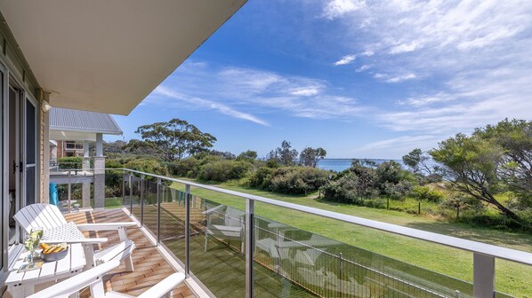 A balcony with white chairs and a table, overlooking a grassy area with trees and a view of the sea. - The Helm, 2/22 Voyager Cl - Stunning waterfront unit with Air Conditioning (Nelson Bay)