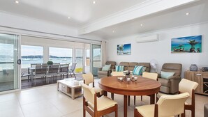 A spacious living area with a round wooden table, comfortable seating, and large windows offering a view of the sea. - Casa Martins, 219 Foreshore Drive - Absolute Waterfront House (Salamander Bay)