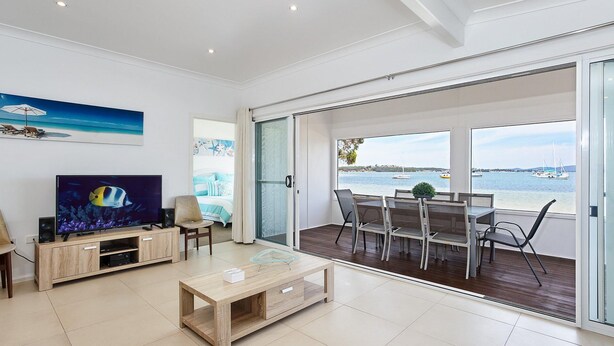 A modern living room with a TV, wooden furniture, and a balcony with a view of the sea. - Casa Martins, 219 Foreshore Drive - Absolute Waterfront House (Salamander Bay)