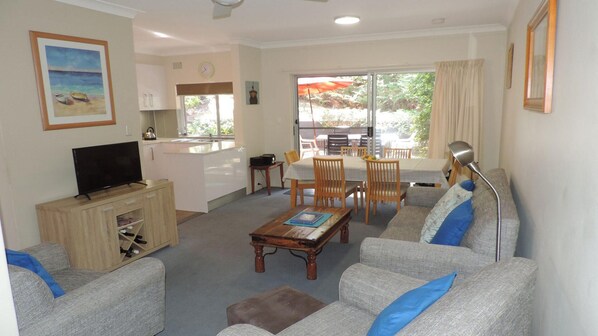 A living room with a sofa, coffee table, and TV. There is a dining area with a table and chairs. A kitchen is visible in the background. - Adriana, 3/83 Ronald Avenue - unit with a backyard (Shoal Bay)