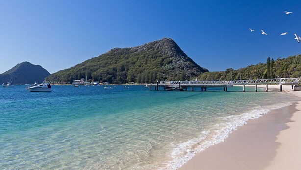 A beach with clear turquoise water, a sandy shore, a pier, and a mountain in the background.