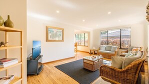 A modern living room with a flat-screen TV, wicker furniture, and a large window offering a view of a building with a tiled roof.