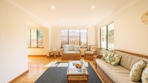 A spacious living room with a sofa, two wicker chairs, a glass coffee table with snacks, and large windows offering a view of trees.