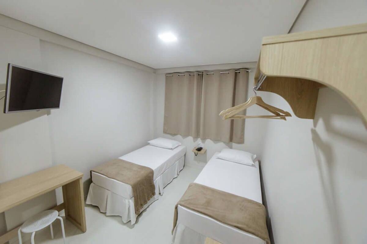 A hotel room with two single beds, a desk, a chair, a TV mounted on the wall, and a wooden wardrobe.