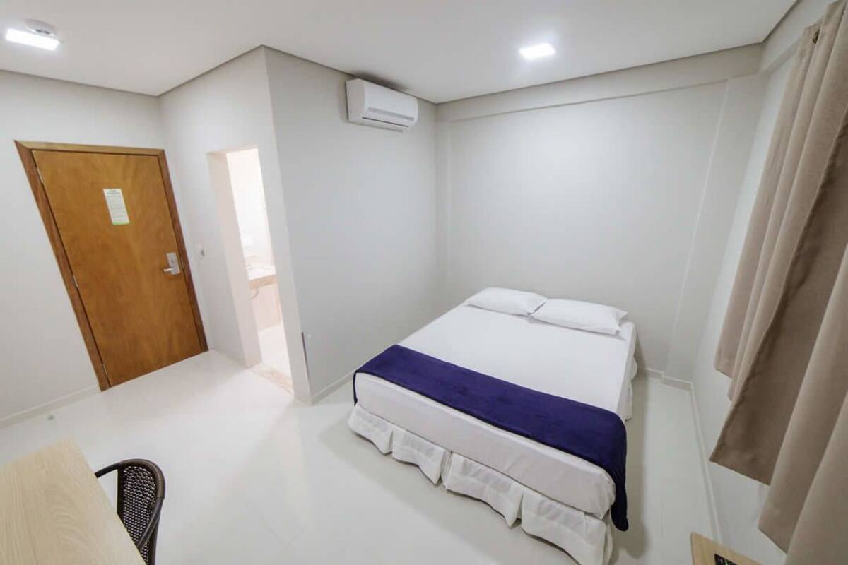 A bedroom with a bed, a chair, a wooden door, an air conditioning unit, and a curtain.
