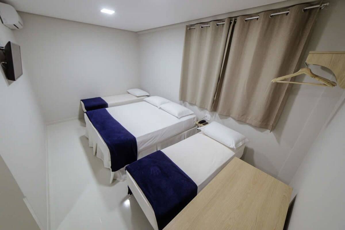 A hotel room with two single beds, a wooden table, a hanging coat rack, and a window with curtains.