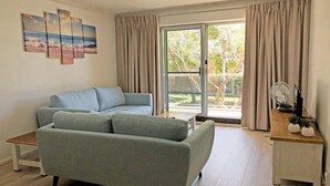 Sala de estar con sofá gris, una mesa de centro de madera y una mesita auxiliar con un ventilador y plantas. Hay una puerta de vidrio corrediza que da a un área exterior con árboles.