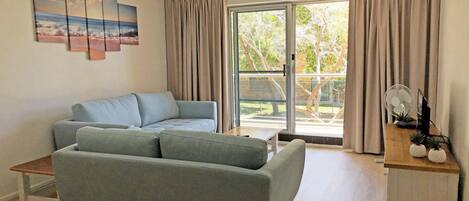A living room with a grey sofa, a wooden coffee table, and a small side table with a fan and plants. There is a sliding glass door leading to an outdoor area with trees.