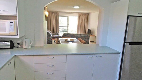 A compact kitchen with a microwave, a kettle, and a refrigerator. There is a dining area with a table and chairs visible through an archway. - Parkview, 10/11-13 Catalina Close - peaceful park views (Nelson Bay)