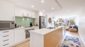 A modern kitchen with white cabinetry, stainless steel appliances, and a central island with a sink. - The Shoal, 104/6-8 Bullecourt Street - linen included & under 200m to beach (Shoal Bay)