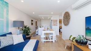 Un salon moderne avec un canapé bleu, un coin repas avec une table blanche et une cuisine en arrière-plan.