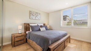 A bedroom with a wooden bed frame, a nightstand, a bedside table, and a window with blinds.