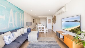 Uma sala de estar moderna com um sofá, uma área de jantar e uma grande TV exibindo uma vista de praia.