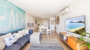 A modern living room with a sofa, a dining area, and a large TV screen displaying a beach view. - The Shoal, 104/6-8 Bullecourt Street - linen included & under 200m to beach (Shoal Bay)