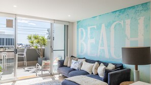 A modern living room with a large sliding glass door leading to a balcony, a blue couch with decorative pillows, and a large wall mural with the word 'BEACH'.