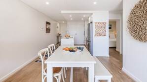 Uma sala de jantar moderna com uma mesa branca, cadeiras e um banco. É visível uma cozinha ao fundo, com uma geladeira e uma pia.