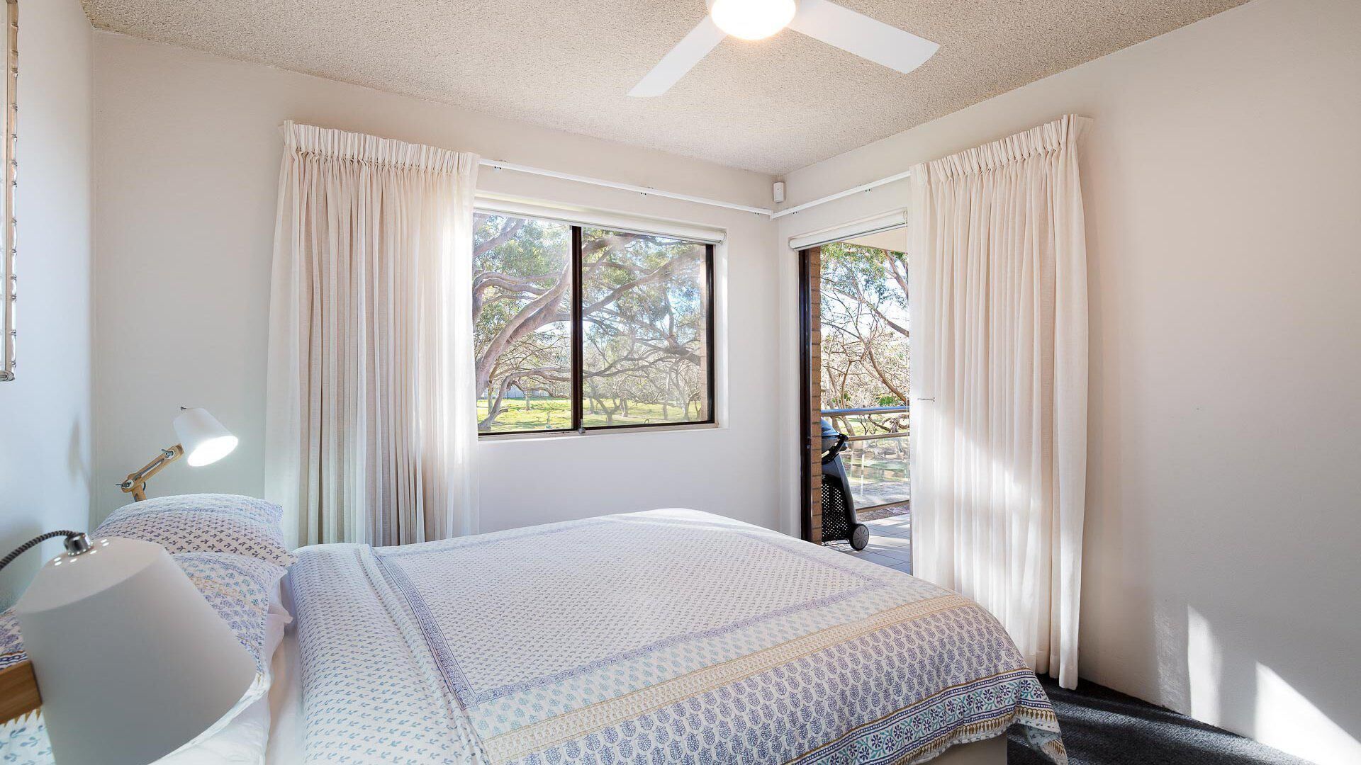 Une chambre à coucher comprenant un lit, un bureau, une chaise, une fenêtre avec des rideaux et un ventilateur de plafond.