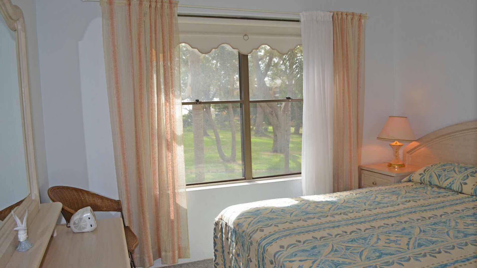 A bedroom with a bed, a bedside table with a lamp, a chair, and a window with curtains.